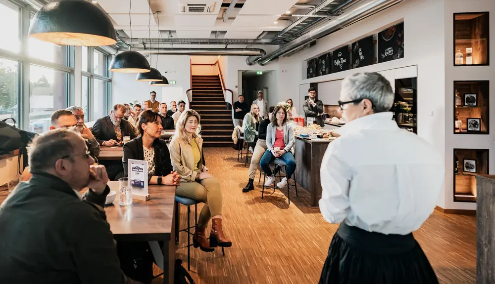 Beim KI-Frühstück im hellen Batix-Café spricht eine Person im weißen Hemd zu den Gästen an Tischen und auf Barhockern